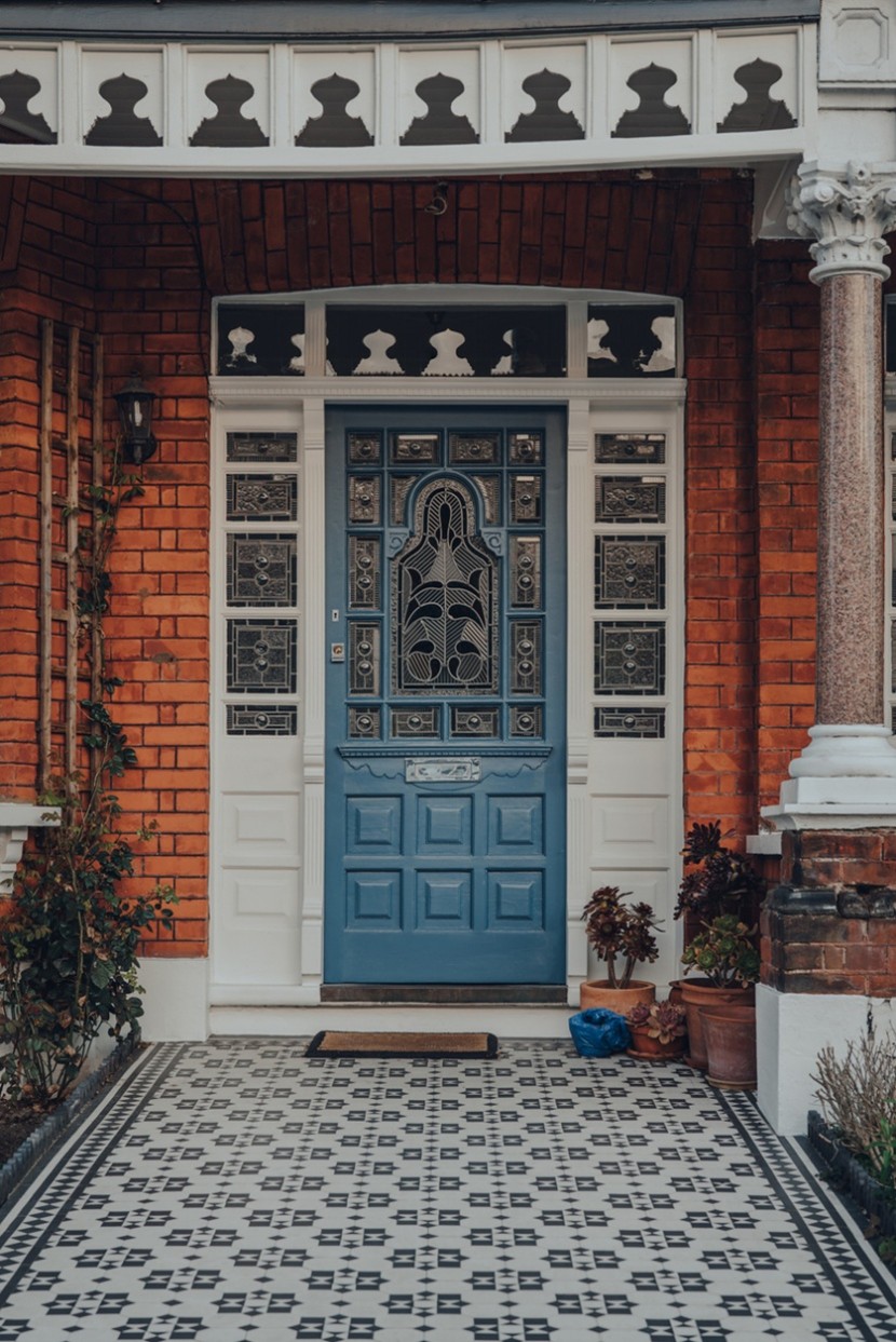 Ornate blue front door with decorative glass panels set in a red brick Victorian-style entrance, framed by white columns and patterned black-and-white tiled flooring.