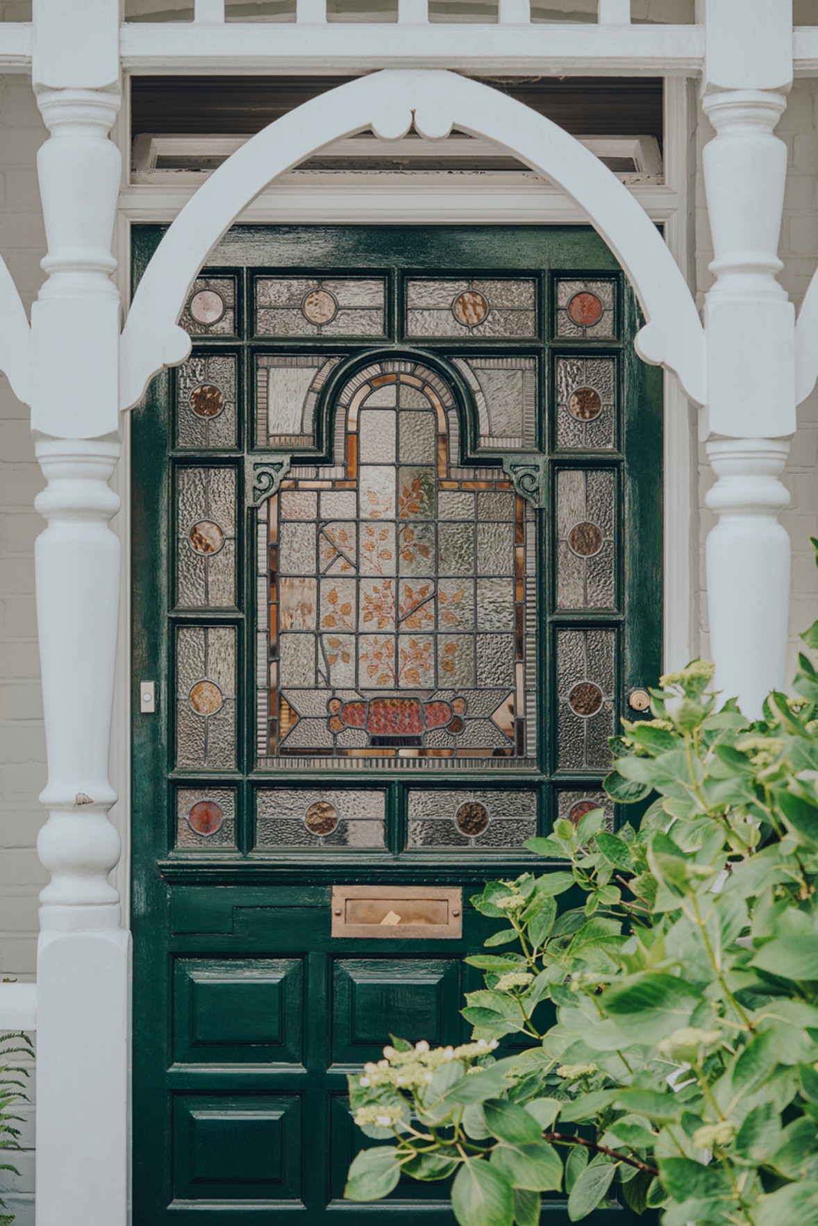 Dark green front door with ornate stained-glass panels, framed by white decorative woodwork and partially surrounded by leafy green plants.