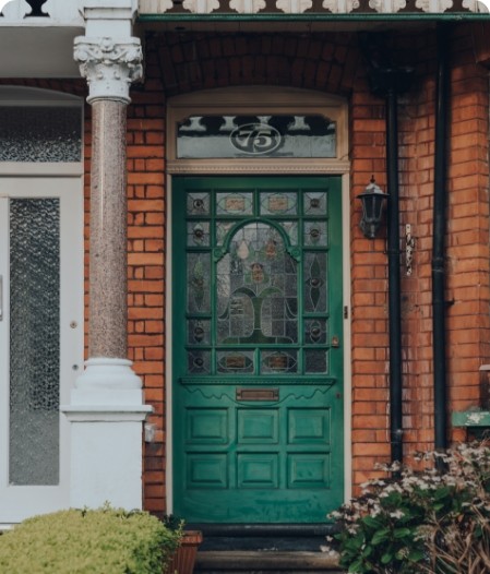 Front entrance of a red brick townhouse with a green stained-glass front door, decorative columns, and a black wall lantern beside the doorway.