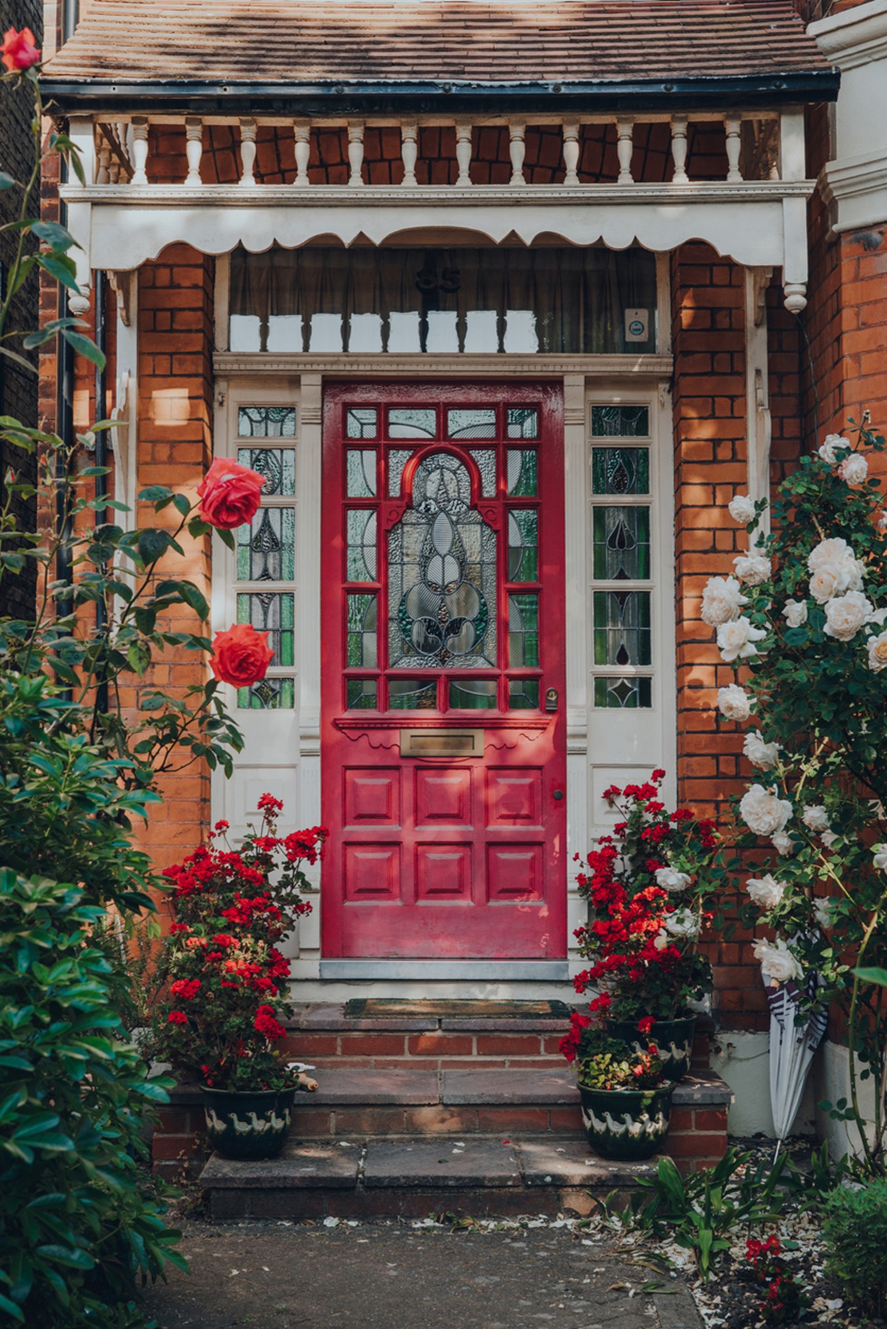 Red front door with stained glass panels on a brick house entrance, surrounded by blooming red and white roses and decorative white trim.