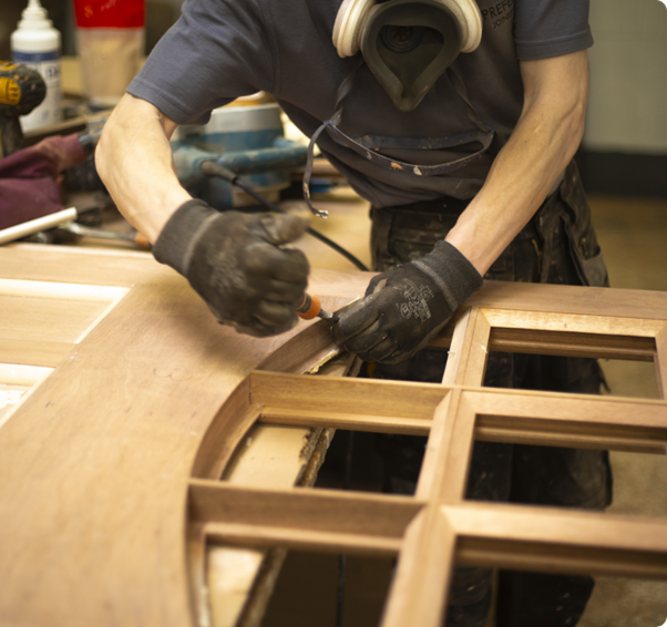 A craftsperson wearing gloves and a respirator mask works on assembling a wooden door with decorative window panes in a workshop.