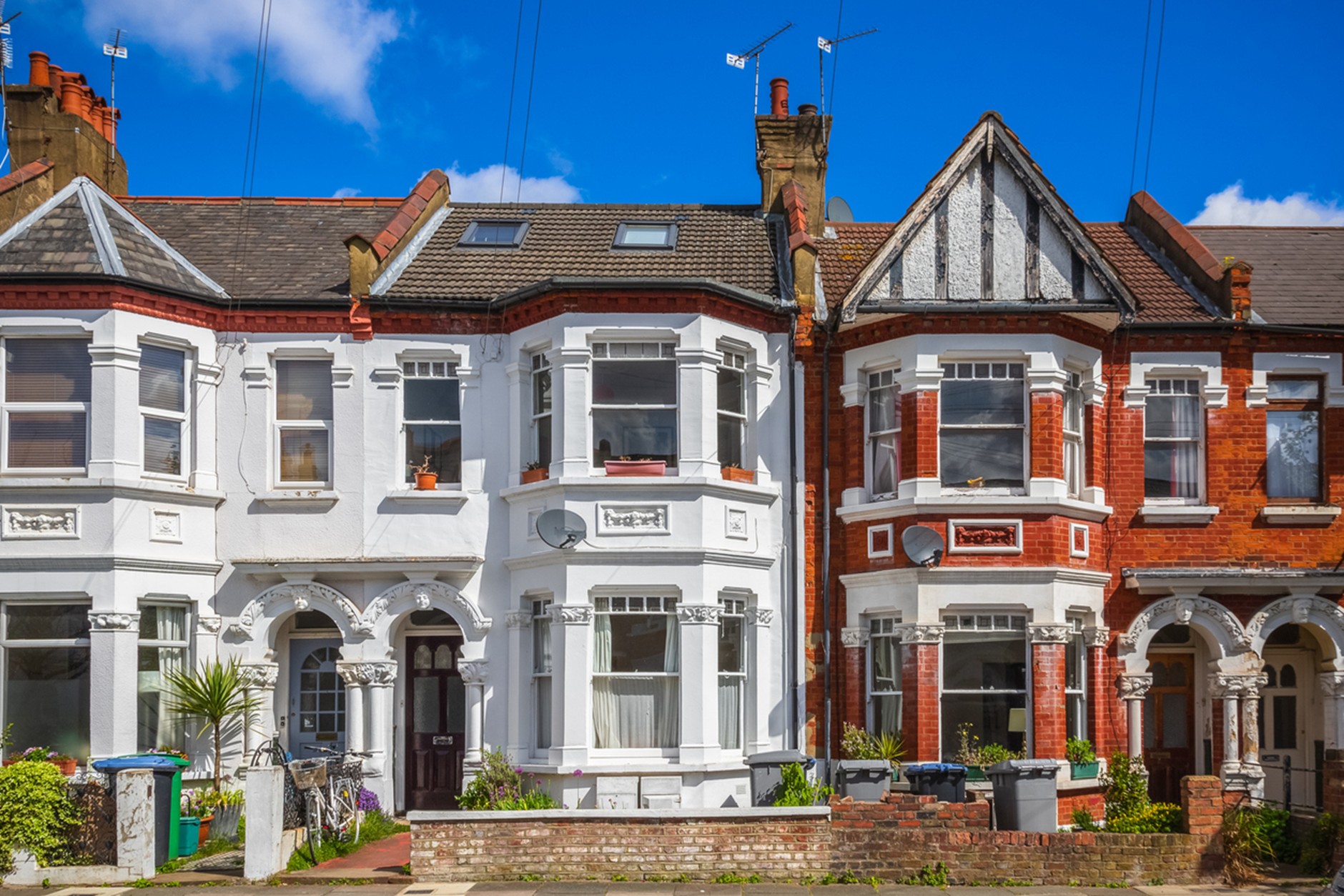 Row of attached Victorian-style townhouses with bay windows and ornate white trim under a bright blue sky, featuring one white-painted home between red brick houses.