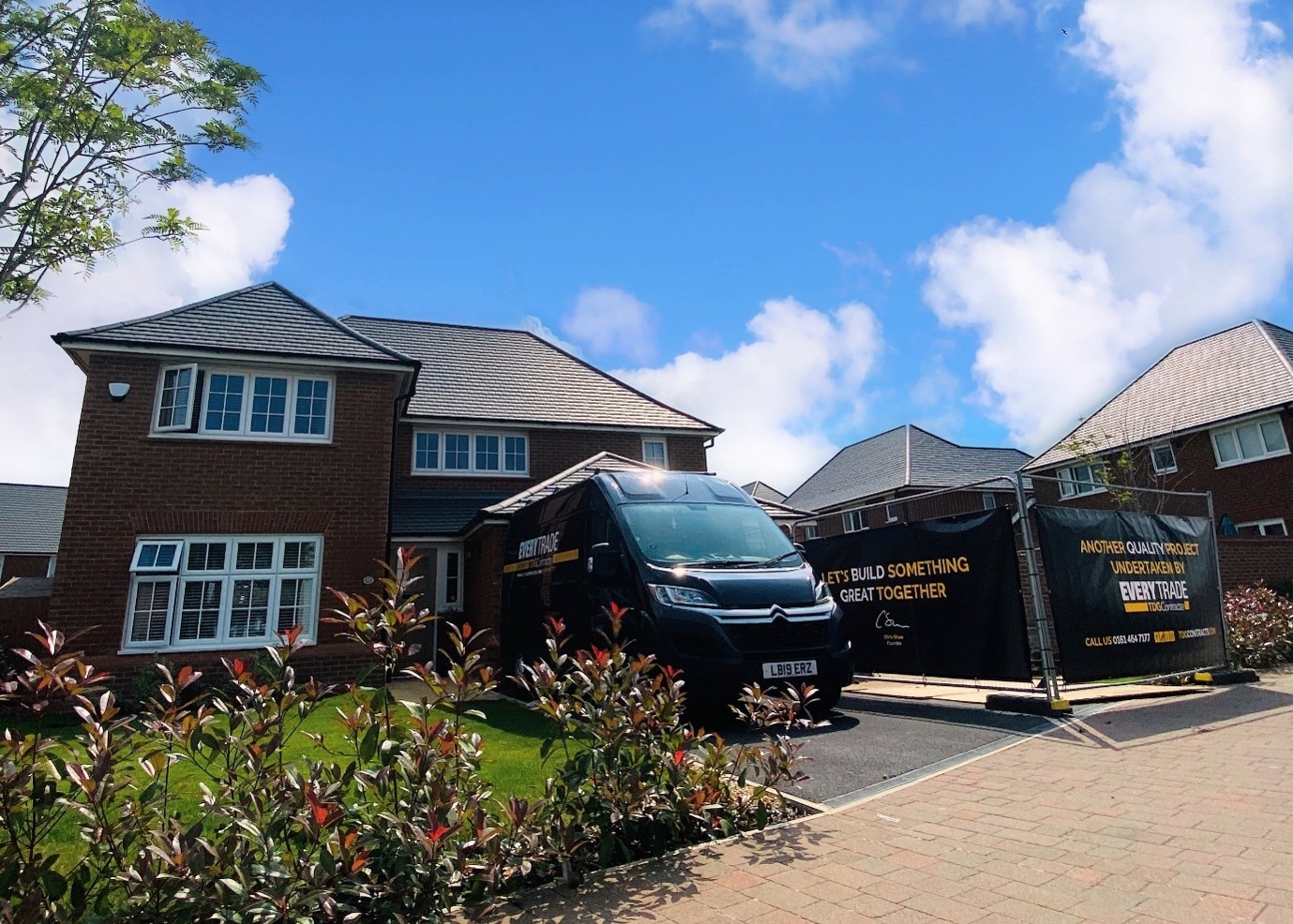 A black contractor van is parked outside a large detached brick home on a sunny day in a modern residential neighborhood. Temporary site fencing and branded construction banners surround part of the property, indicating an active home renovation or building project. Decorative shrubs line the foreground, while blue skies and scattered clouds provide a bright backdrop.