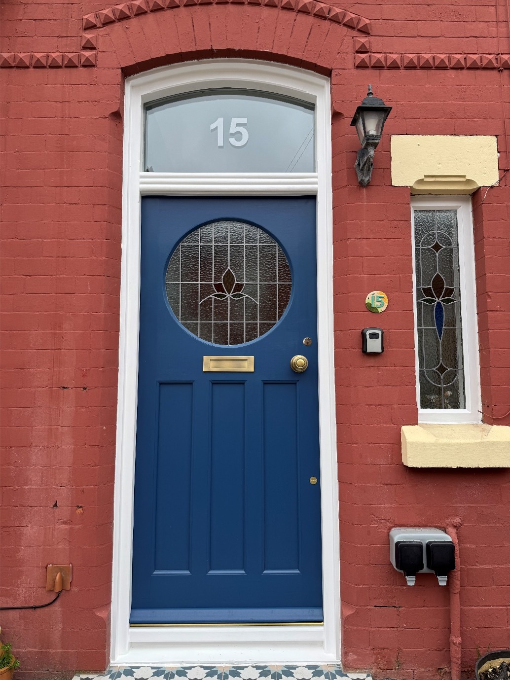 A blue front door with a circular decorative stained-glass window is set within a red brick terraced house. White trim frames the doorway, with the number "15" displayed in the arched transom window above. A black wall lantern, brass letterbox and door knob, and a narrow stained-glass side window add traditional character to the entrance.