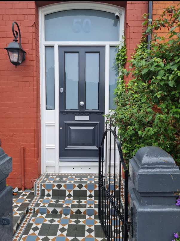 A dark blue front door with frosted glass panels sits within a red brick facade, framed by white trim. Patterned geometric tiles lead up the entrance, with a black metal gate and lush green climbing plants bordering the walkway. The house number "50" is visible in the frosted transom window above the door.