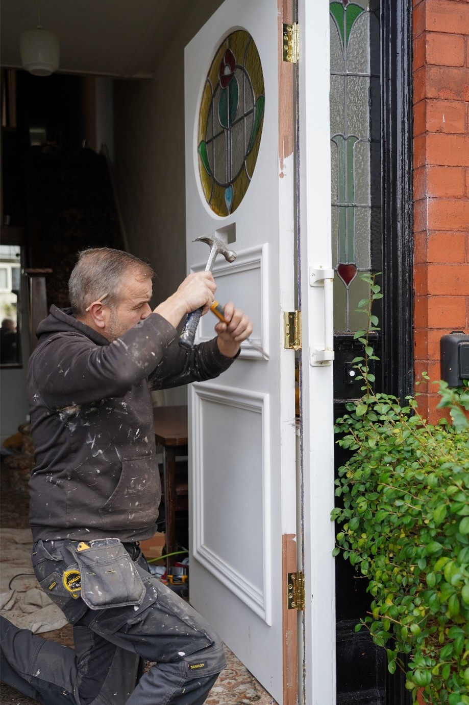 A tradesperson kneels in the doorway of a red brick house, using a hammer to fit or adjust a white front door. The door features a circular stained-glass panel with green, yellow, blue, and red details, while matching stained-glass sidelights frame the entrance. Paint marks on the worker’s clothing and exposed wood along the door edge suggest ongoing renovation work. A leafy shrub grows beside the doorway, and the interior hallway is visible in the background.