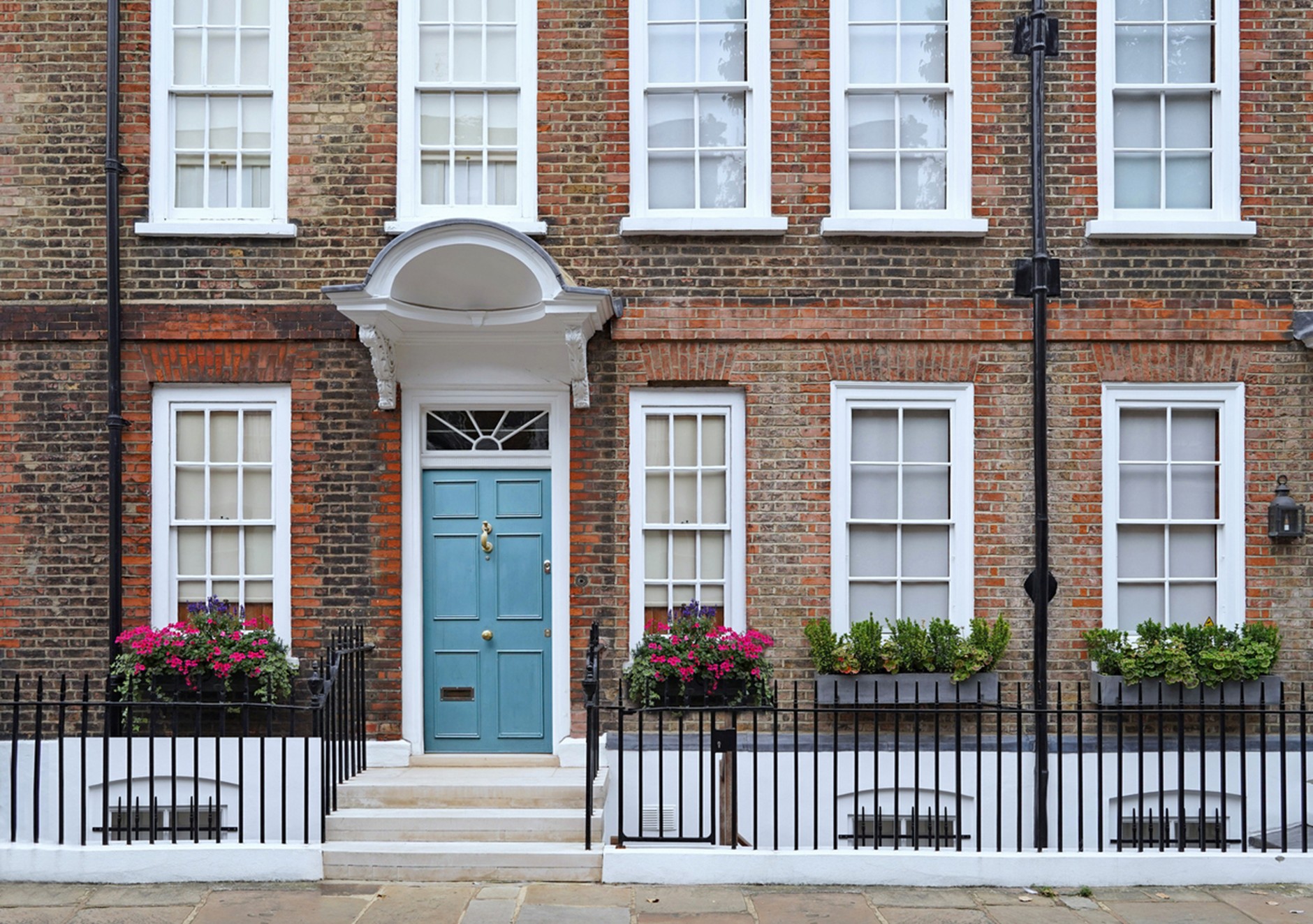 Brick townhouse façade with a pale blue front door beneath a white arched portico, flanked by tall sash windows, flower boxes filled with pink blooms and greenery, black iron railings, and a short set of stone steps leading up from the sidewalk.