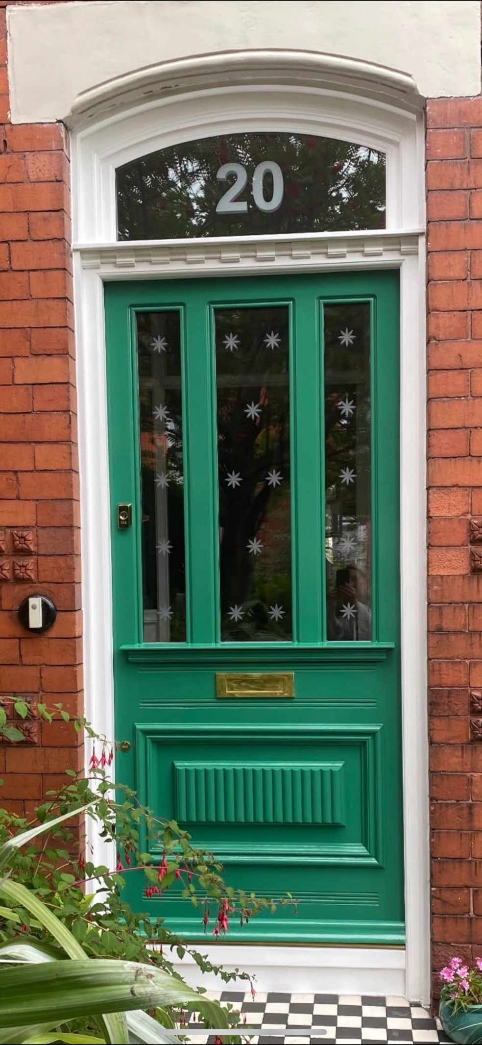 Bright emerald-green front door set within a red brick townhouse facade. The door features three tall glass panels decorated with white starburst motifs, a brass mail slot, and white trim. An arched transom window above displays the number 20. Green plants and pink flowers frame the entrance, with a black-and-white checkerboard doorstep below.