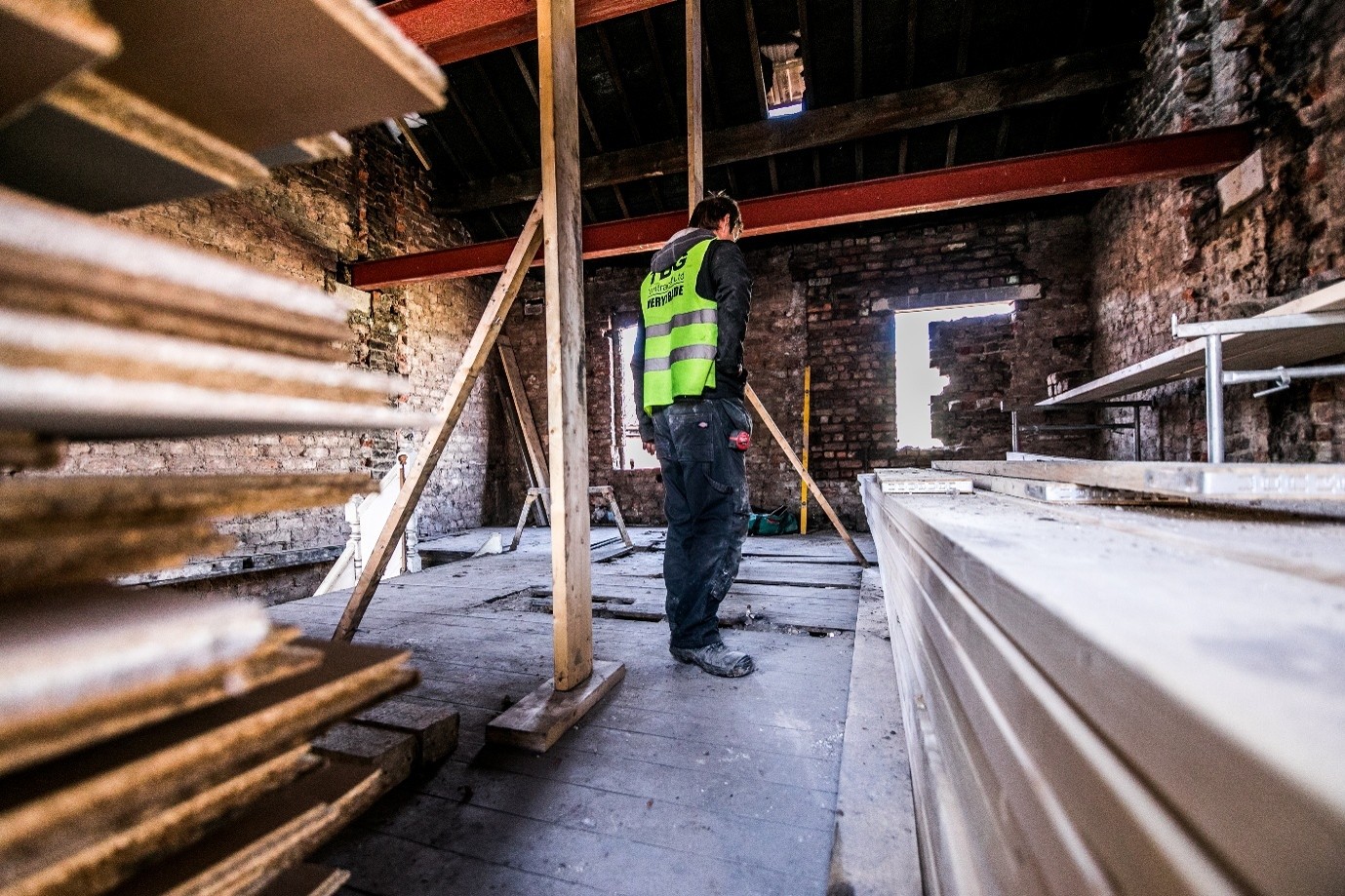 Construction worker wearing a high-visibility vest stands inside a partially renovated brick building, surrounded by stacks of lumber and drywall, with exposed beams and temporary wooden supports visible throughout the unfinished interior.
