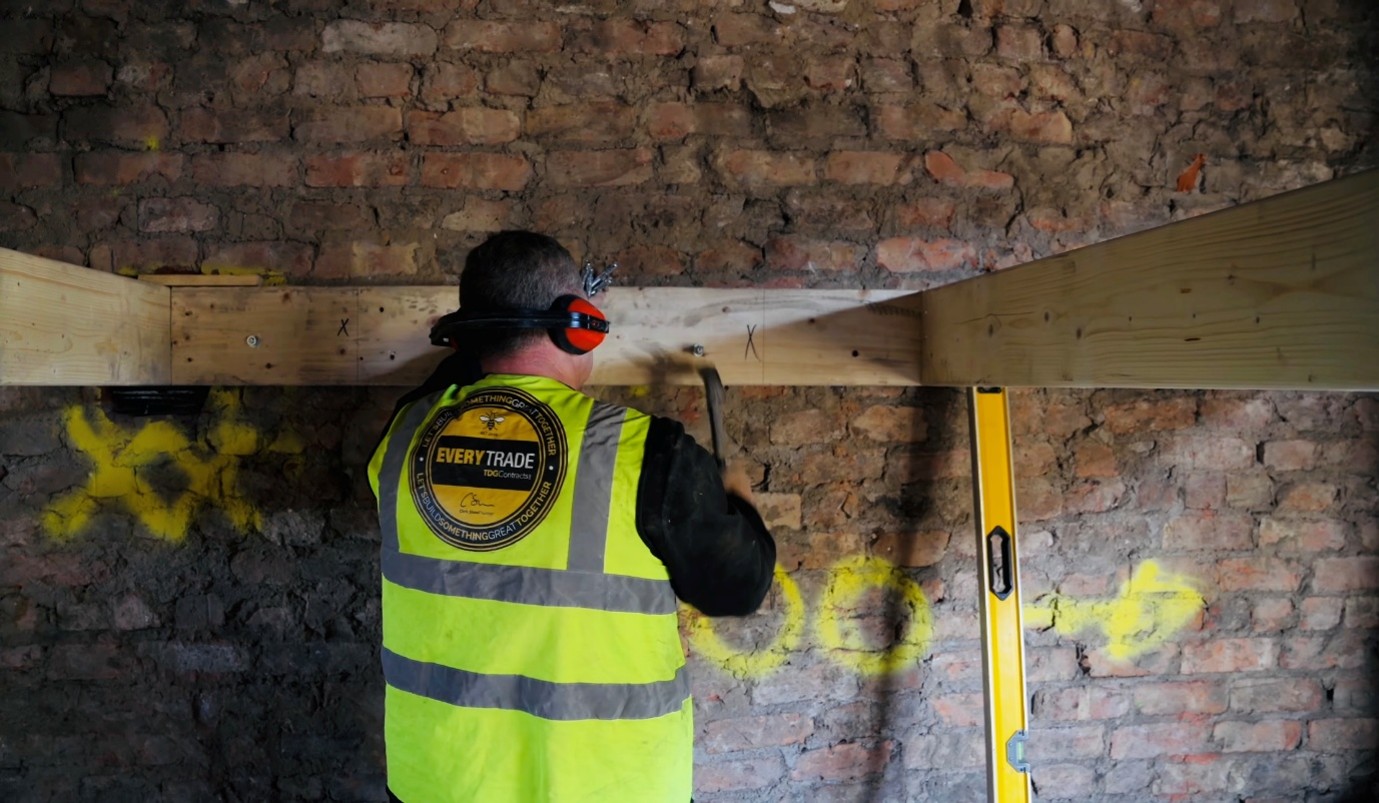 Construction worker wearing a high-visibility vest and hearing protection secures a timber beam to an exposed brick wall, using a hammer while a spirit level stands nearby.