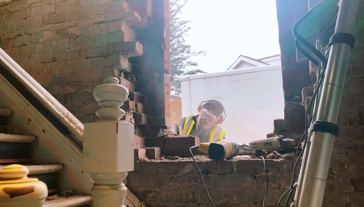 Construction worker wearing safety goggles, ear protection, and a high-visibility vest removes bricks from a damaged brick wall inside an older building. A staircase with a wooden banister is visible on the left, while power tools and renovation equipment sit on the partially demolished wall. Bright daylight shines through the large opening being created in the wall.