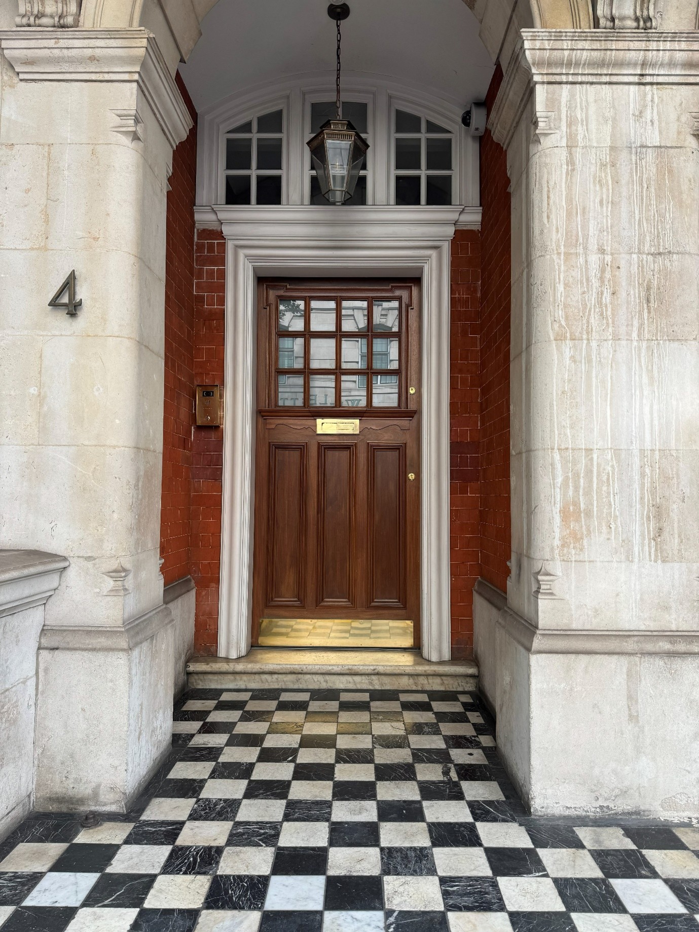 Elegant townhouse entrance with a dark wooden front door framed by white stone columns and red brick walls, featuring a hanging lantern overhead and a black-and-white checkerboard marble floor leading to the doorway.
