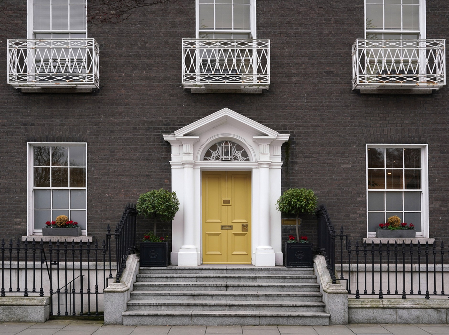 Front entrance of a symmetrical brick townhouse with a pale yellow double door framed by white columns and a decorative pediment, reached by a short flight of stone steps. Black wrought-iron railings and potted shrubs flank the entrance, with large sash windows and three small white balconies above.
