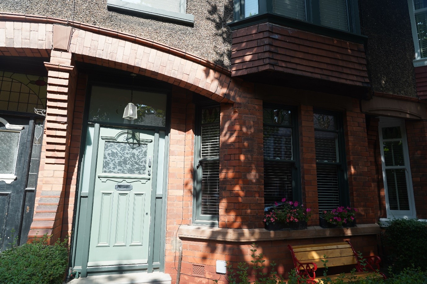 Front view of a red-brick terraced house with a pale green front door beneath a brick archway, dark-framed bay windows, flower boxes on the windowsill, and a wooden bench in the small front garden.