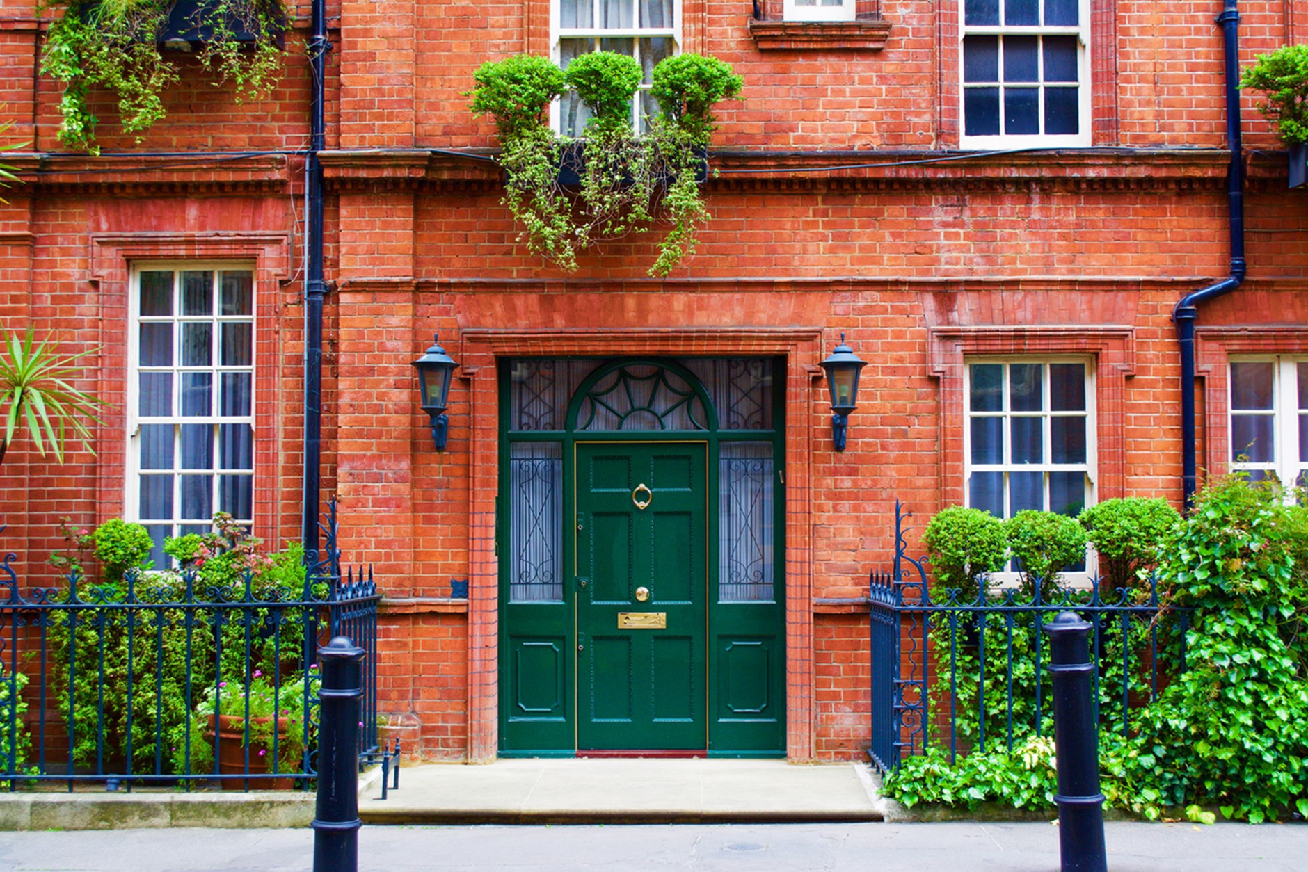 Red brick townhouse façade with a dark green front door framed by decorative glass panels and black lantern-style wall lights. Tall sash windows, neatly trimmed shrubs, hanging planters, and black wrought-iron railings create a symmetrical and well-maintained entrance.