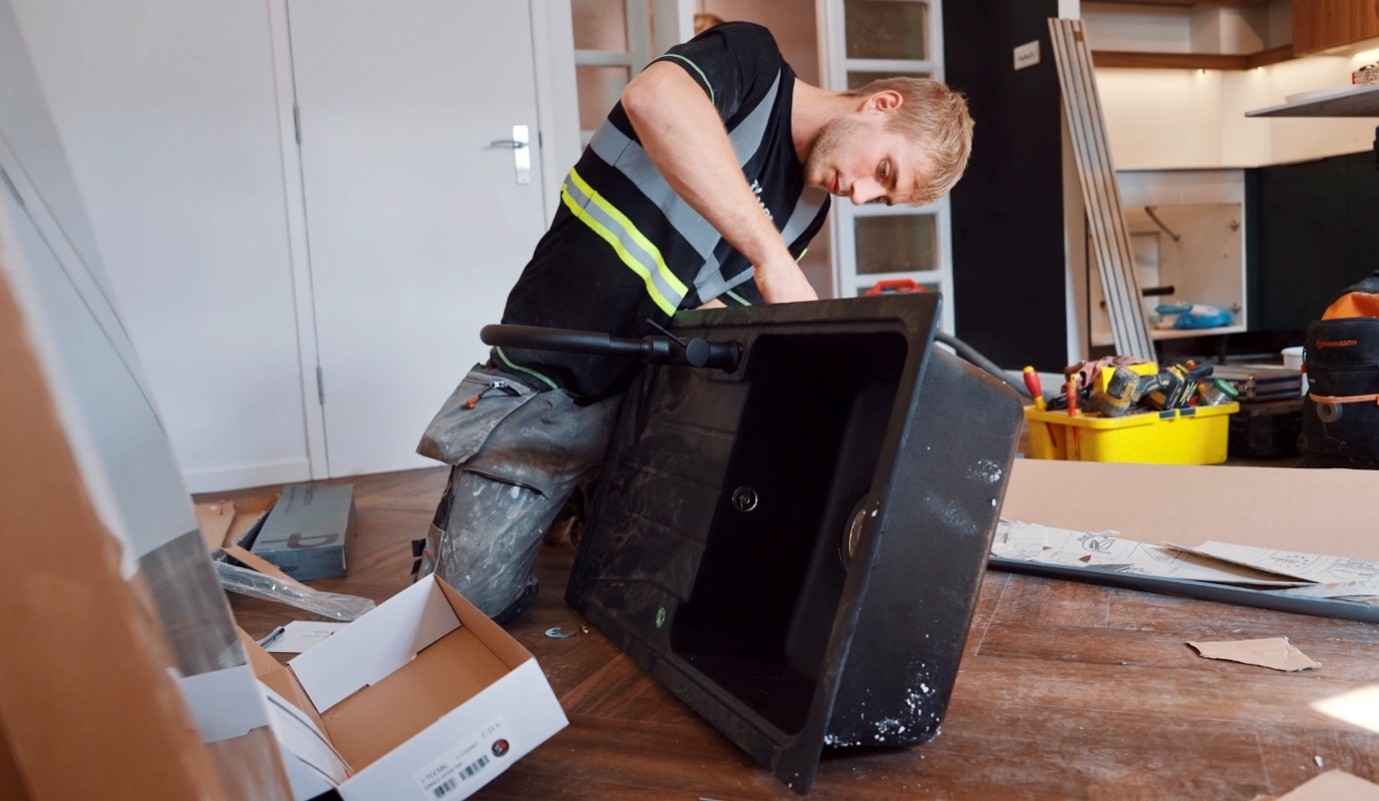 Tradesperson wearing a high-visibility vest kneels on the floor of a partially renovated kitchen, installing a black sink while tools, packaging, and construction materials are spread around the workspace.