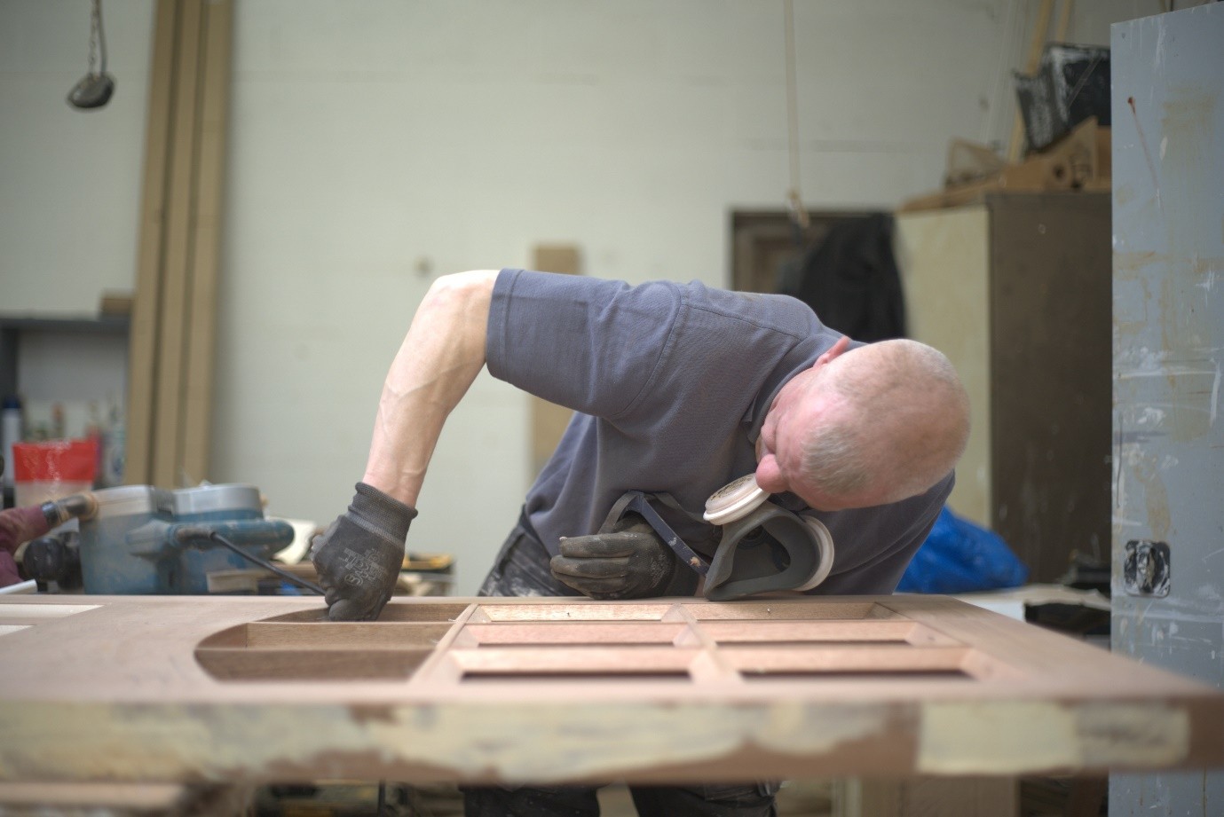 A craftsperson wearing protective gloves and a respirator mask sands a wooden door in a workshop. The door lies flat on a workbench, showing multiple recessed panels and a curved opening on one side. Tools, timber boards, and workshop equipment are visible in the background.