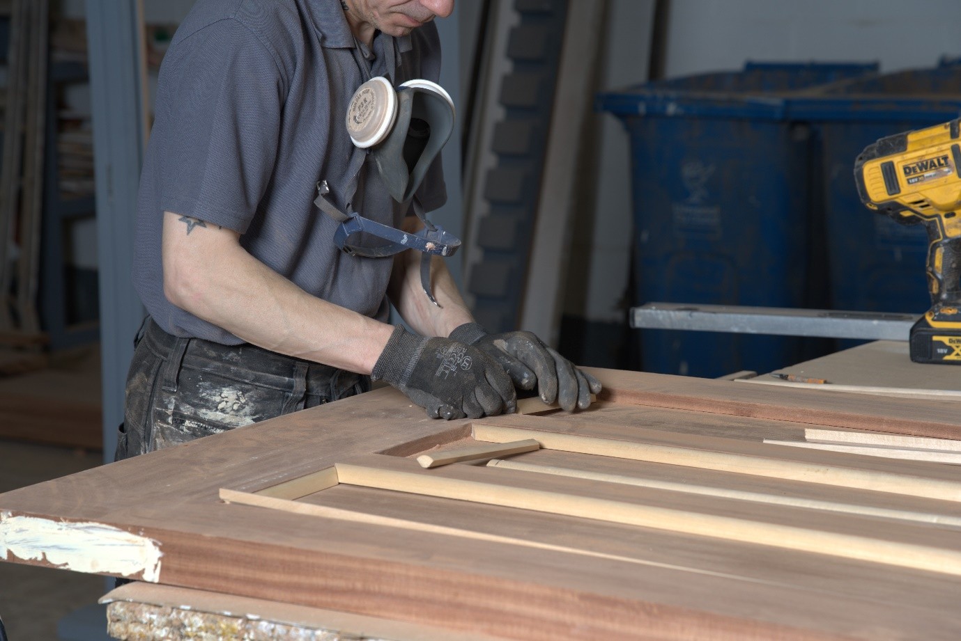 A workshop craftsperson wearing protective gloves and a respirator mask works on a wooden door laid flat on a workbench. The door frame and timber panels are being assembled or adjusted by hand, with woodworking tools and equipment visible in the workshop background.