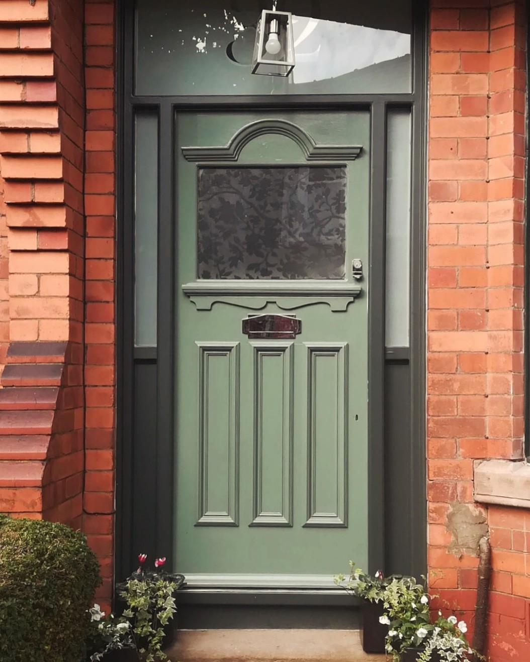 Decorative sage green front door set within a red brick house entrance. The door features raised period-style mouldings, a frosted glazed panel with a floral pattern, and a central letterbox. Dark trim surrounds the doorway, with a glazed transom window above and a hanging lantern light. Small potted plants and flowers sit at the base of the entrance.