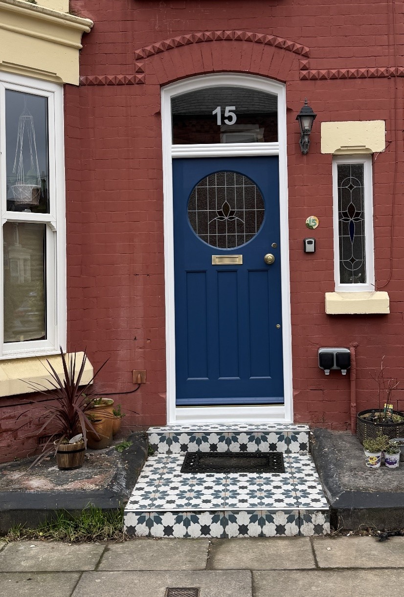 Front entrance of a red brick terraced house with a blue front door numbered 15. The door features a circular stained-glass panel, framed by white trim, with patterned black-and-white tiled steps leading up to it. Narrow decorative windows flank the entrance, and potted plants sit on either side of the doorway.